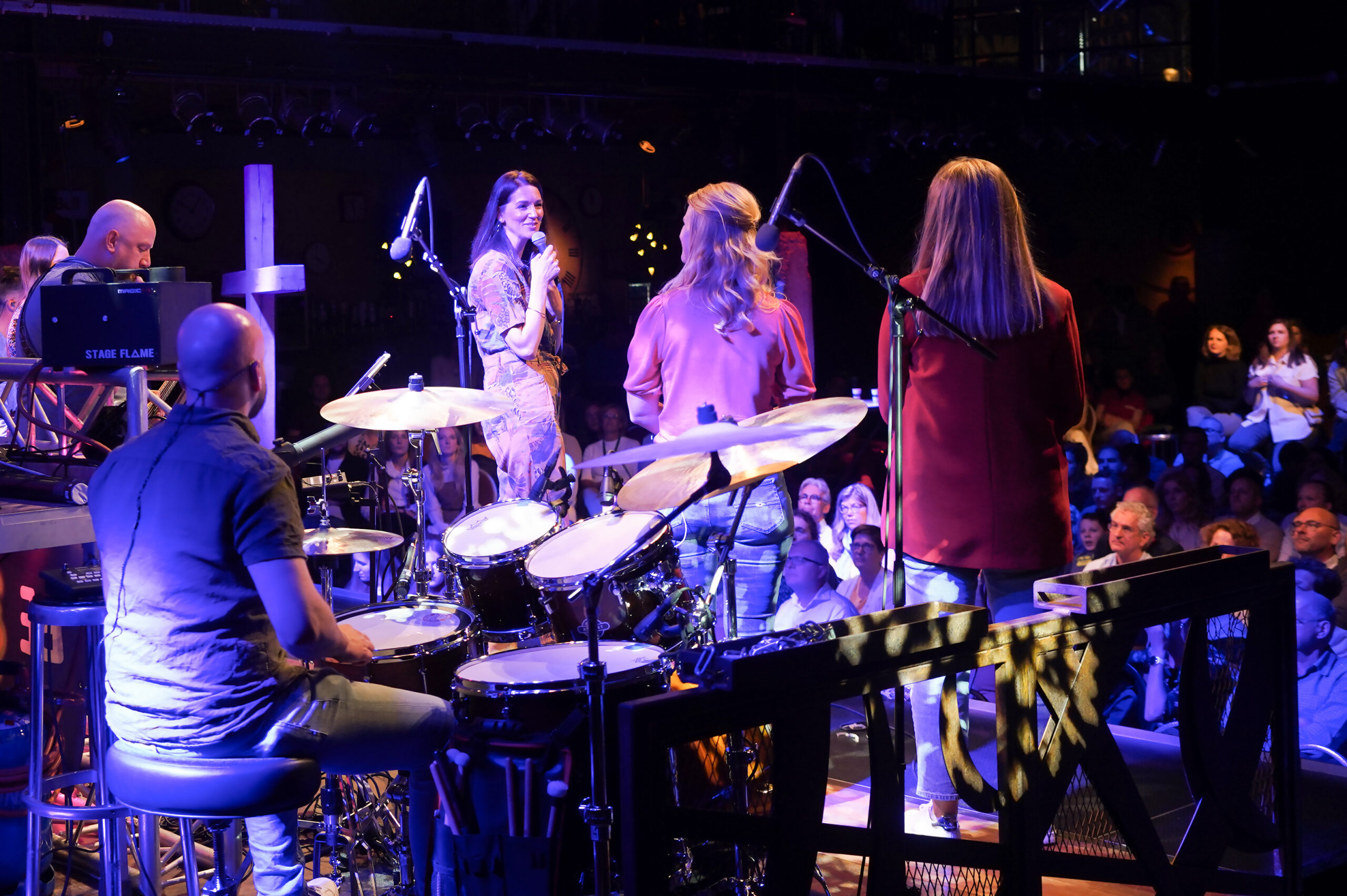 Drie zangeressen bij de microfoon en een drummer op het podium tijdens een dienst van Mozaiek0548