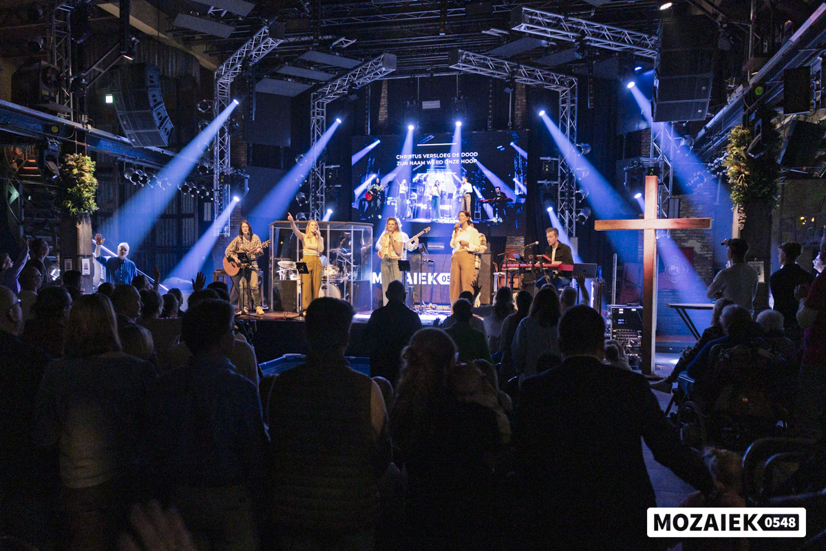 Overzicht van de zaal met podium, band en houten kruis tijdens de doopdienst tijdens de doopdienst van Mozaiek0548