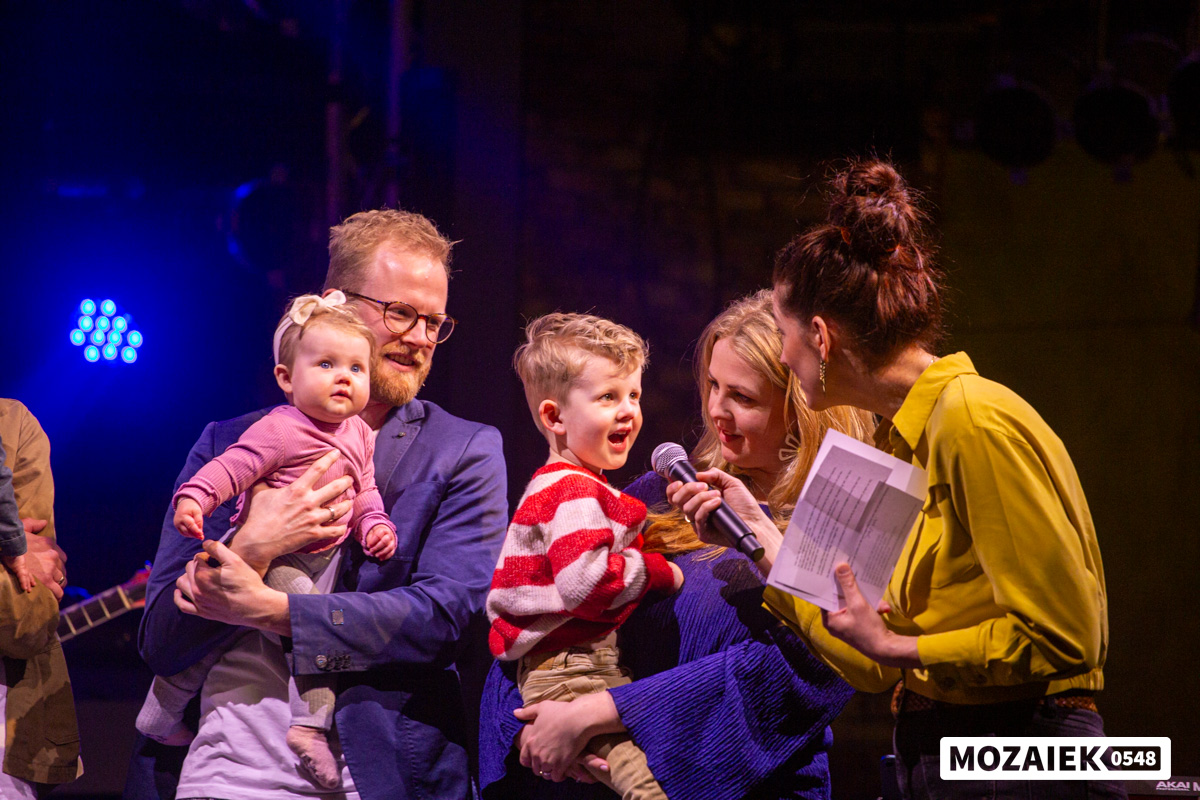Een jongen vertelt iets in de microfoon naast zijn ouders tijdens een opdraagdienst van Mozaiek0548