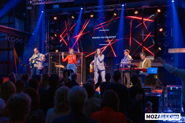 De band speelt op het podium met de liedtekst op het scherm en bezoekers op de voorgrond