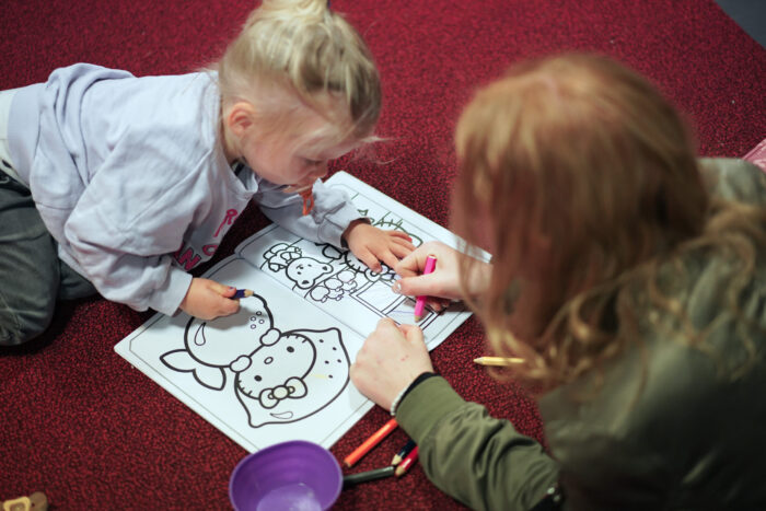 Een peuter maakt samen met een vrijwilliger een kleurplaat op de vloer bij Mozaiek Kids