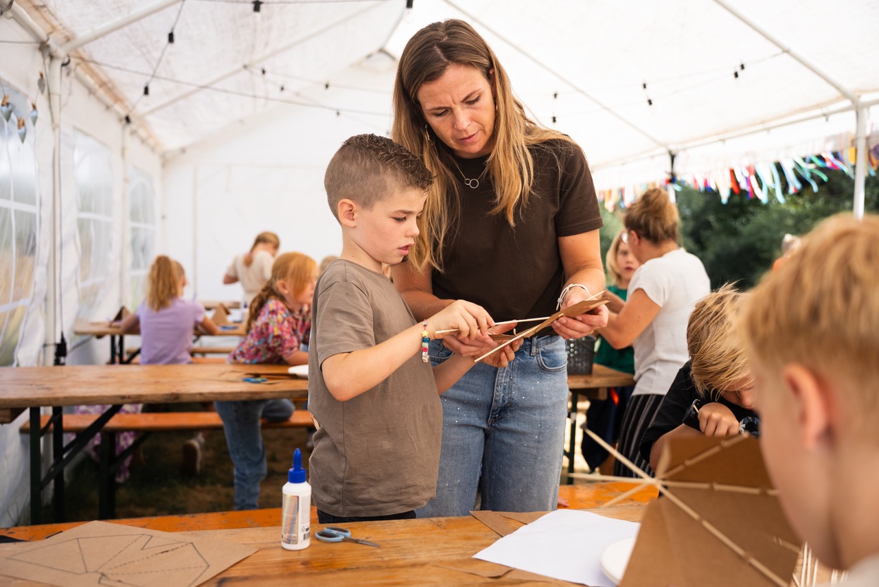 Begeleidster helpt een jongen met knutselen aan de tafel in de tent tijdens de Kidsdag van Mozaiek0548