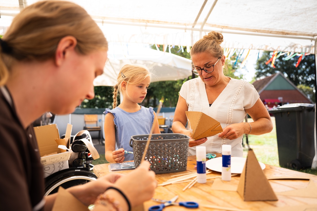 Kinderen en begeleiders knutselen aan een lange tafel onder de tent tijdens de Kidsdag van Mozaiek0548