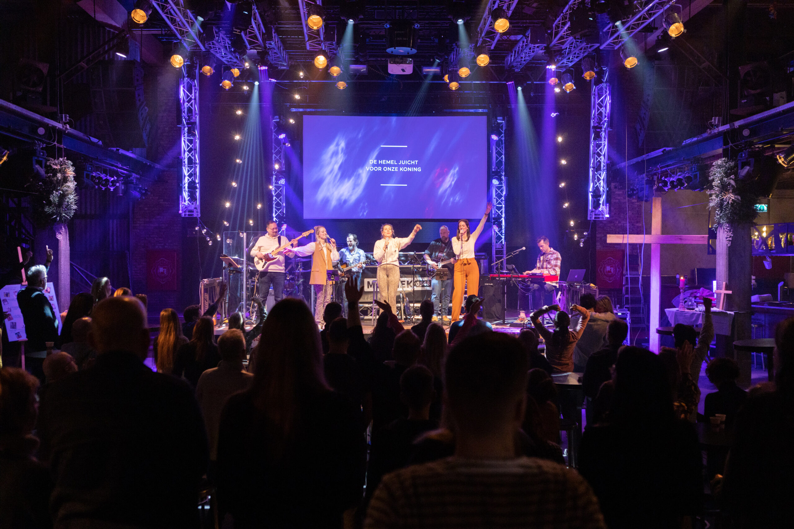Zaaloverzicht dienst Mozaiek0548 met worshipband en de songtekst De hemel juicht voor onze Koning op het scherm