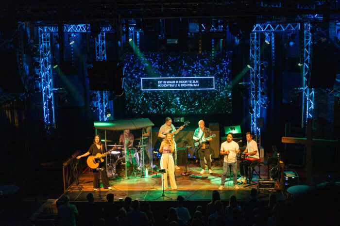 Worshipband op het podium van Mozaiek0548 met blauwe lichtshow en Nederlandse songtekst op het LED-scherm