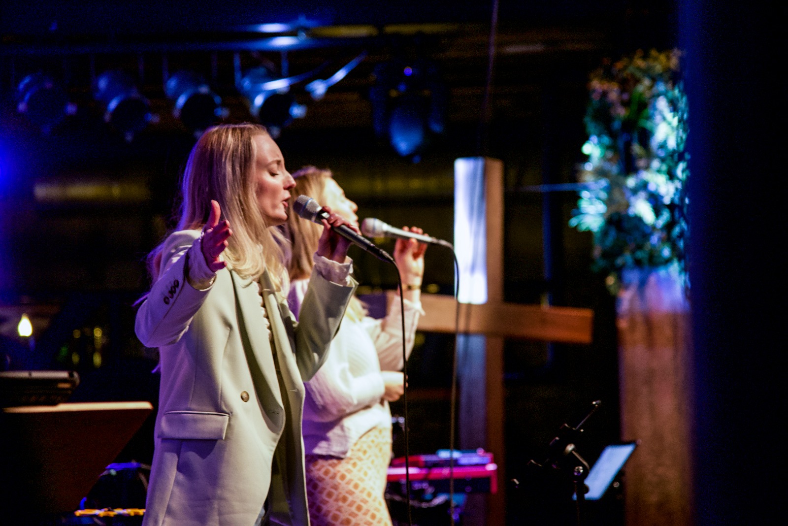Zangeres zingt met microfoon tijdens de worship in Mozaiek0548, met een houten kruis op de achtergrond