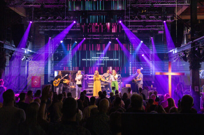 Zaaloverzicht van een dienst in Mozaiek0548 met paarse lichtshow en de songtekst Hosanna op het scherm