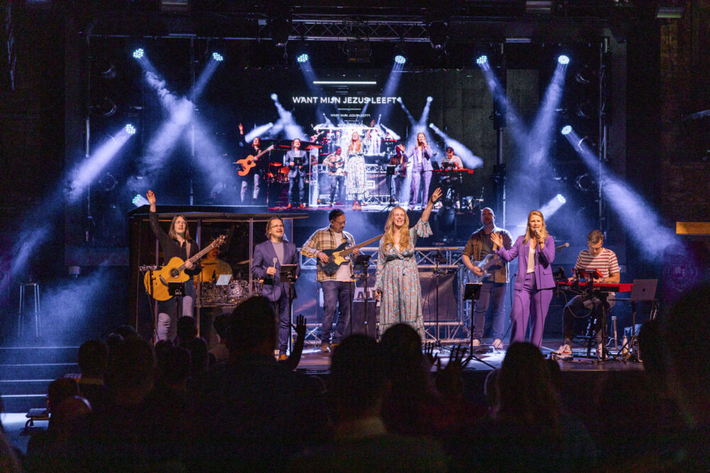 Worshipband van Mozaiek0548 op het podium met lichtstralen en de tekst Want mijn Jezus leeft op het scherm