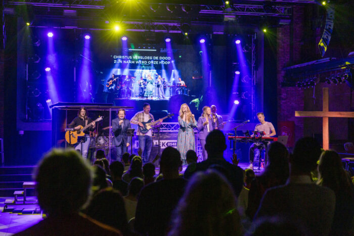 Worshipband van Mozaiek0548 op het podium met blauwe lichtshow en de songtekst Christus versloeg de dood